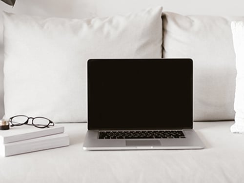 Modern laptop with blank display and stack of books with glasses lying on comfortable couch in stylish room
